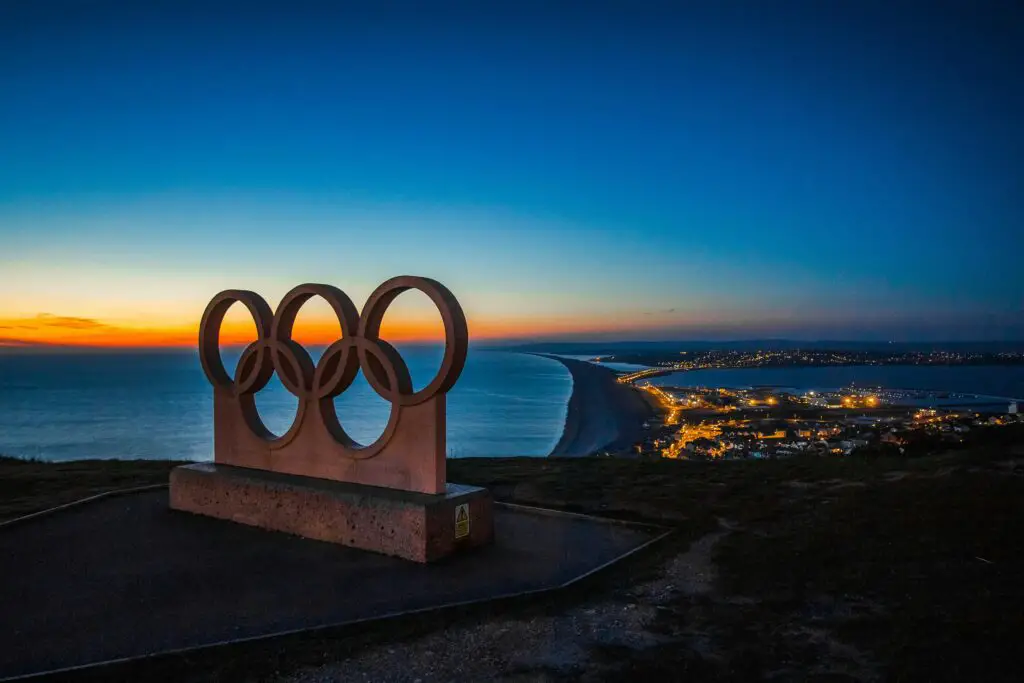 Scenic sunset view of the Olympic rings monument by the sea with city lights in the distance.